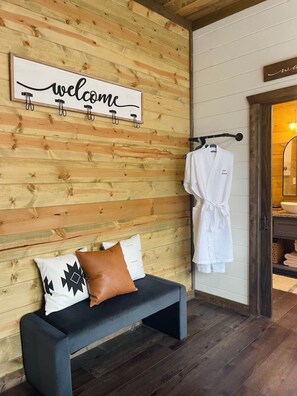 Een houten wand met een 'welcome'-bord, een bank met kussens en een badjas hangend aan een rek.