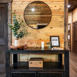 Un mur en bois orné d’un miroir rond, une pancarte de bienvenue et une petite boîte en bois posée sur une console noire.