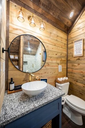Een houten badkamer in een chalet, met een ronde spiegel, een granieten aanrecht, een witte wastafel, een toilet en een klein plankje met toiletartikelen.