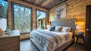 A bedroom with a large bed, wooden walls, a window, and a view of trees. - Luxury Cabin! Hot tub, Pet Friendly, Foosball! (Broken Bow)