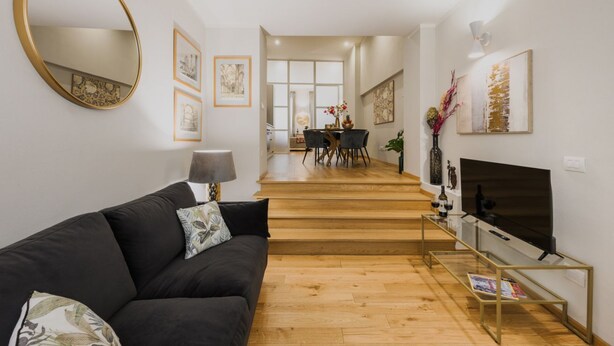 Ein modernes Wohnzimmer mit einem schwarzen Sofa, einem Glastisch, einem Fernseher und einer Treppe, die in einen Essbereich führt.