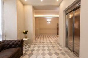Un couloir d’hôtel avec un sol à carreaux, un canapé, une petite table sur laquelle est posé un vase de fleurs et un ascenseur.