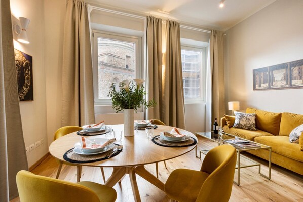 A dining area with a round table, yellow chairs, a vase with flowers, and a view of buildings through large windows.