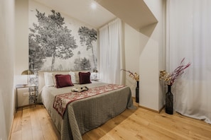 Un dormitorio con una cama, una mesita de noche, una lámpara, un muro con un mural de un árbol y dos jarrones con ramas.