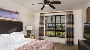 A hotel room with a bed, a ceiling fan, a large window with curtains, a television, and a painting on the wall.