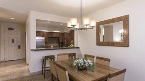 A dining area with a wooden table and chairs, a chandelier, a kitchen in the background, and a large mirror on the wall.