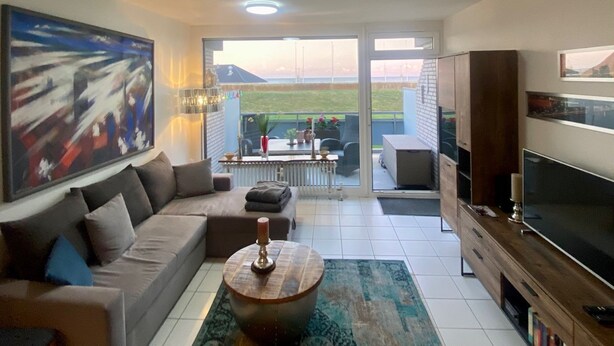Una sala de estar con sofá, mesa de centro, televisor y un cuadro grande. Hay un balcón con vistas al exterior.