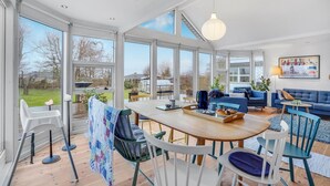 A spacious living area with a large wooden table, blue sofas, and a view of the outdoors through large windows.