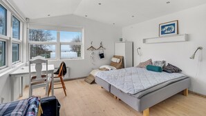 A bedroom with a bed, a desk, and a chair. There are windows, a wall-mounted shelf, and a hanging light fixture.
