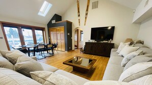 Ein großzügiges Wohnzimmer mit einer großen Couch, einem Couchtisch und einem Fernseher. Es gibt einen Essbereich mit Tisch und Stühlen sowie eine Küchenzeile mit Schränken und einem Herd.