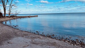 Ein Kiesstrand mit einer Holzsteganlage, die sich in ruhiges Wasser erstreckt, umgeben von kahl stehenden Bäumen unter blauem Himmel.