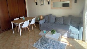 Ein Wohnzimmer mit grauem Sofa, einem Holzesstisch mit weißen Stühlen, einem kleinen weißen Beistelltisch mit Pflanze und einem gemusterten Teppich.