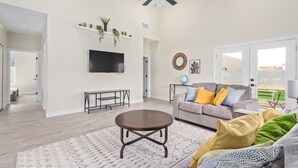 Un salón espacioso con televisor de pantalla plana, un sofá gris con cojines coloridos, una mesa de centro de madera y un ventilador de techo.