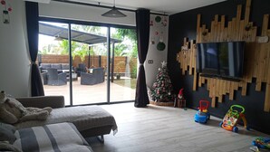 Ein modernes Wohnzimmer mit großem Fernseher, bequemen Sitzgelegenheiten, einem Weihnachtsbaum und Blick auf eine Terrasse.