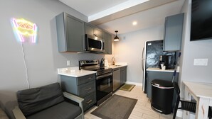 A compact kitchen with grey cabinets, a microwave, stove, and a small refrigerator. There is a black trash bin and a black chair near the counter. - Ski in ski out! Schuss Mountain. Sleeps 4 WIFI, POOL, full kitchen.  (Mancelona)