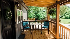 A wooden deck with a table and chairs, a black door, and a view of greenery. - New Tiny Lake Home (Salt Lick)