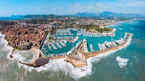 Vue aérienne d’une ville côtière avec un port de plaisance rempli de bateaux, un fort historique et une zone urbaine densément construite, avec des montagnes en arrière-plan.