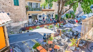 Un café en plein air, avec des tables et des chaises, entouré d’immeubles et d’un arbre.