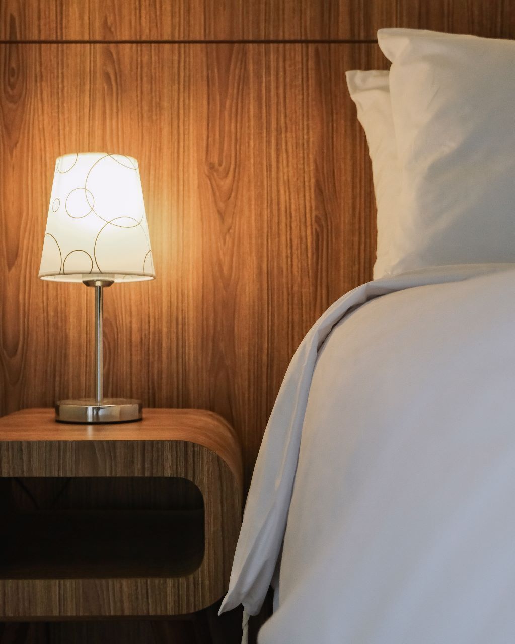 A wooden nightstand with a lamp and a bed with white bedding.