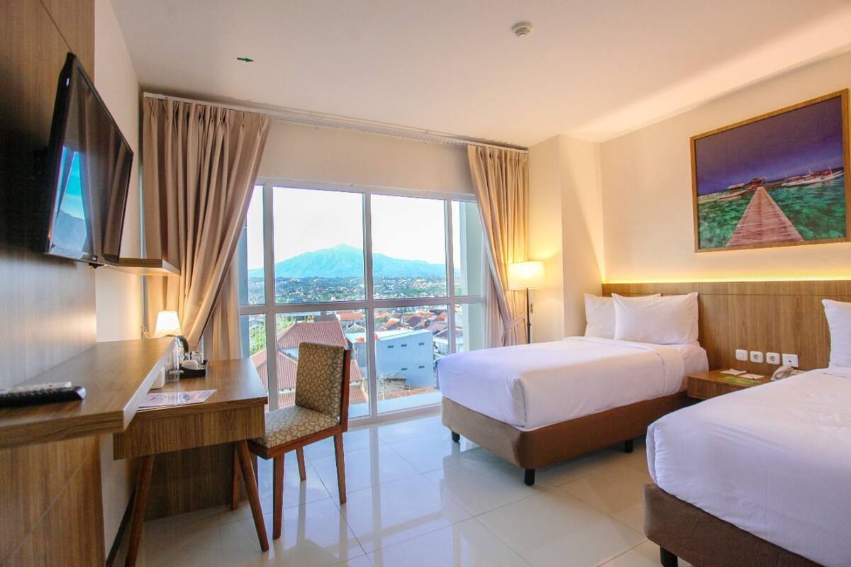 A hotel room with two beds, a desk, a chair, a TV, and a large window with a view of a mountain and cityscape.