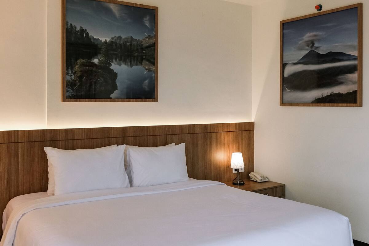 A hotel room with a large bed, two white pillows, a wooden headboard, a bedside table with a lamp, and two framed landscape paintings on the walls.