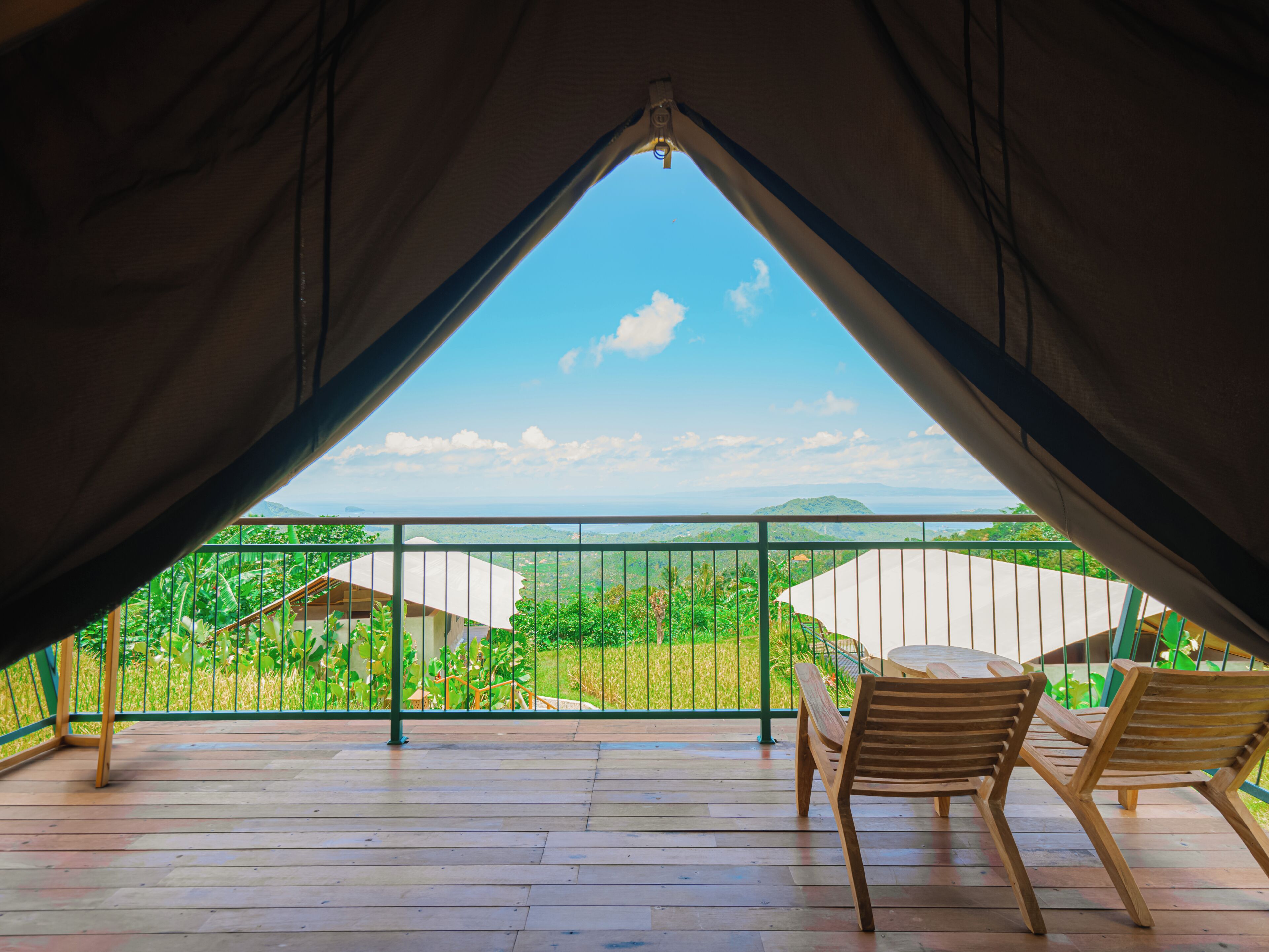 Vista interior de una carpa con cubierta de madera, dos sillas de madera y un paisaje verde visible a través de una baranda metálica.