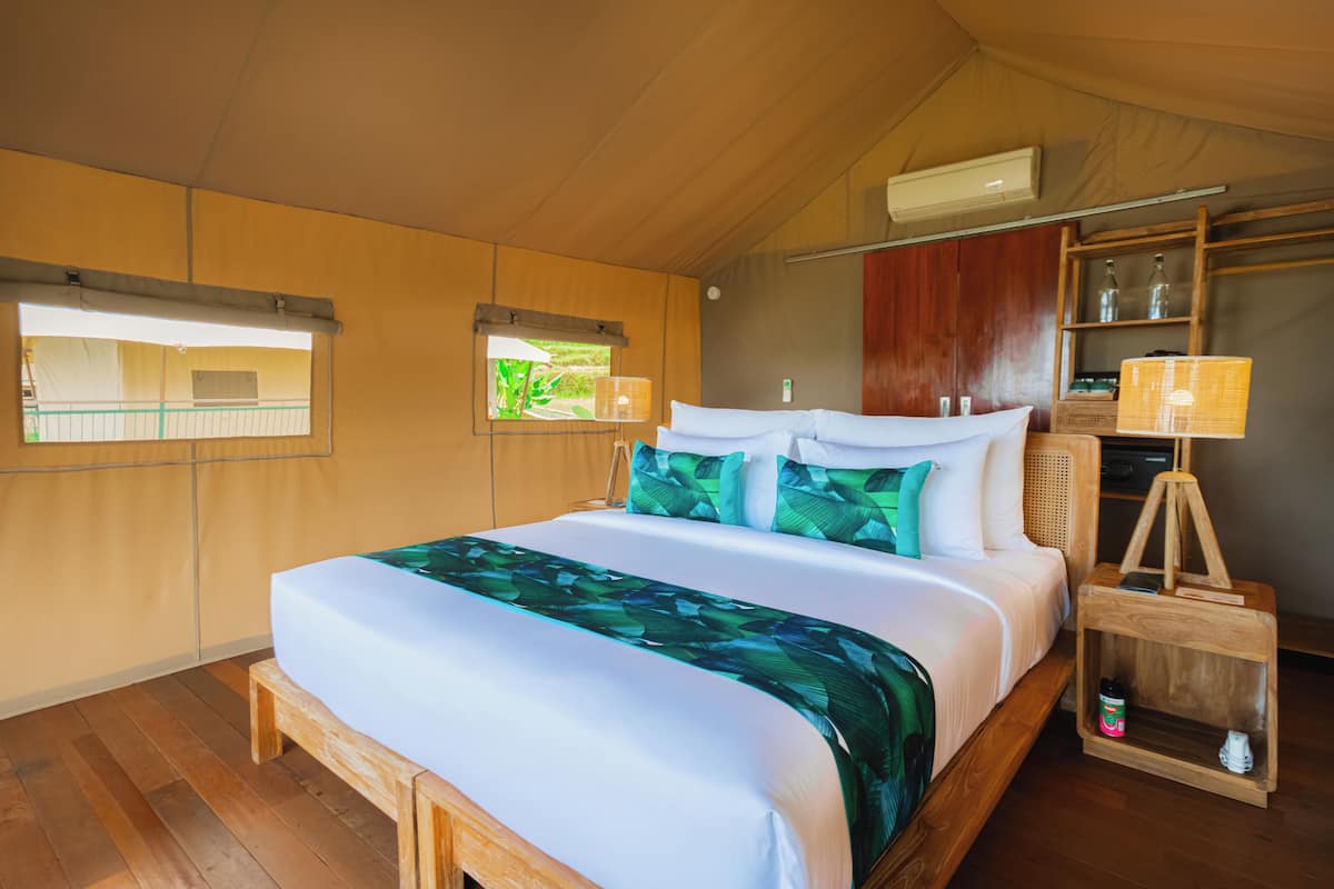 Uma cama de madeira com colcha branca e verde, uma luminária de cabeceira e uma pequena mesa de cabeceira com uma garrafa.