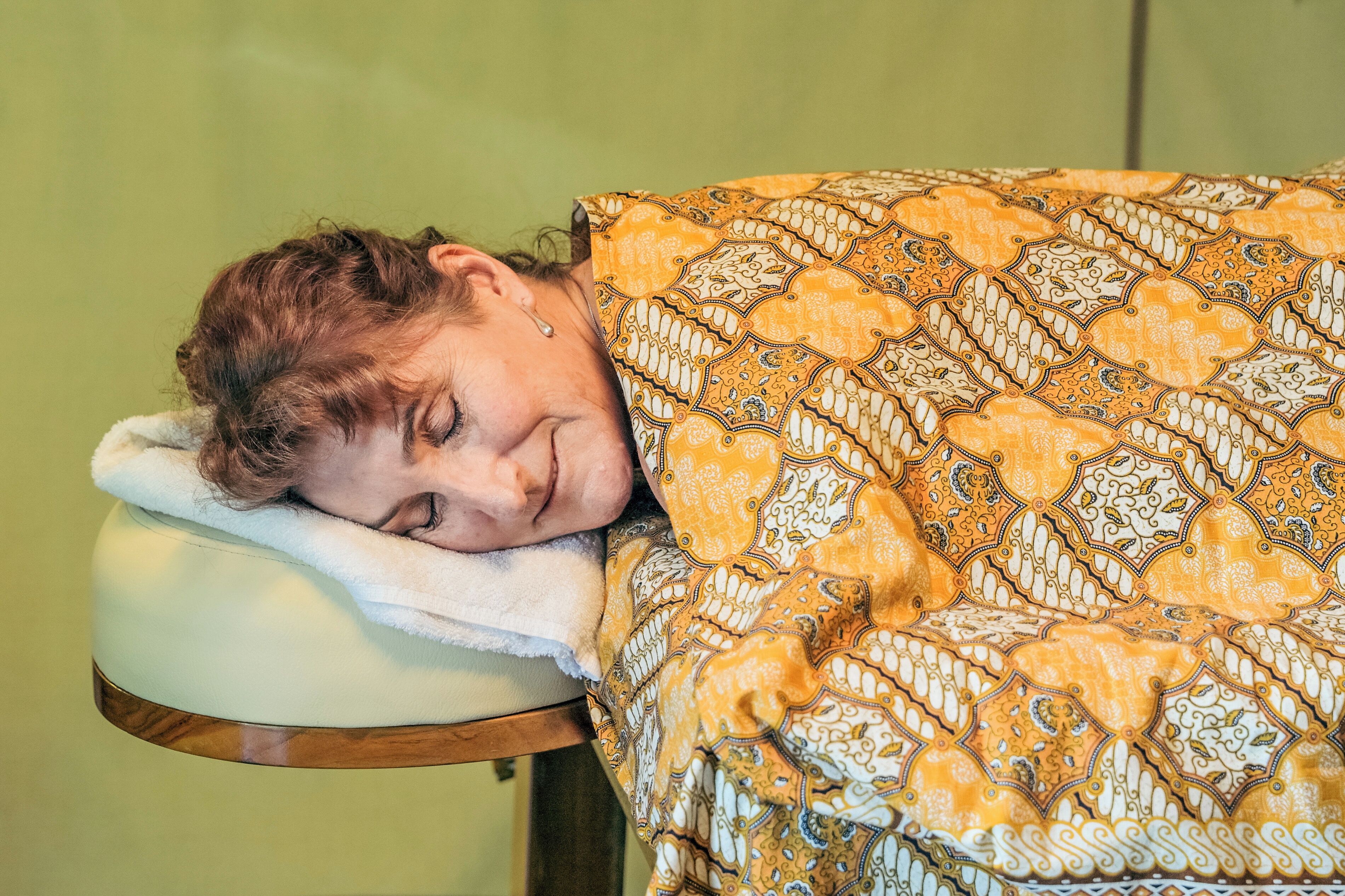 A person resting on a white headrest in a room with a patterned blanket.