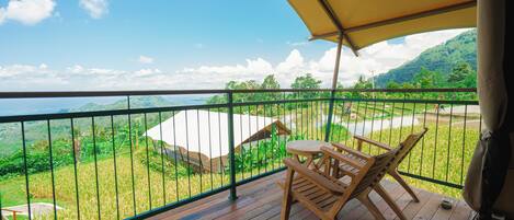 Una terraza de madera con una mesa, sillas, un paraguas grande y vistas a un paisaje verde con una tienda blanca.