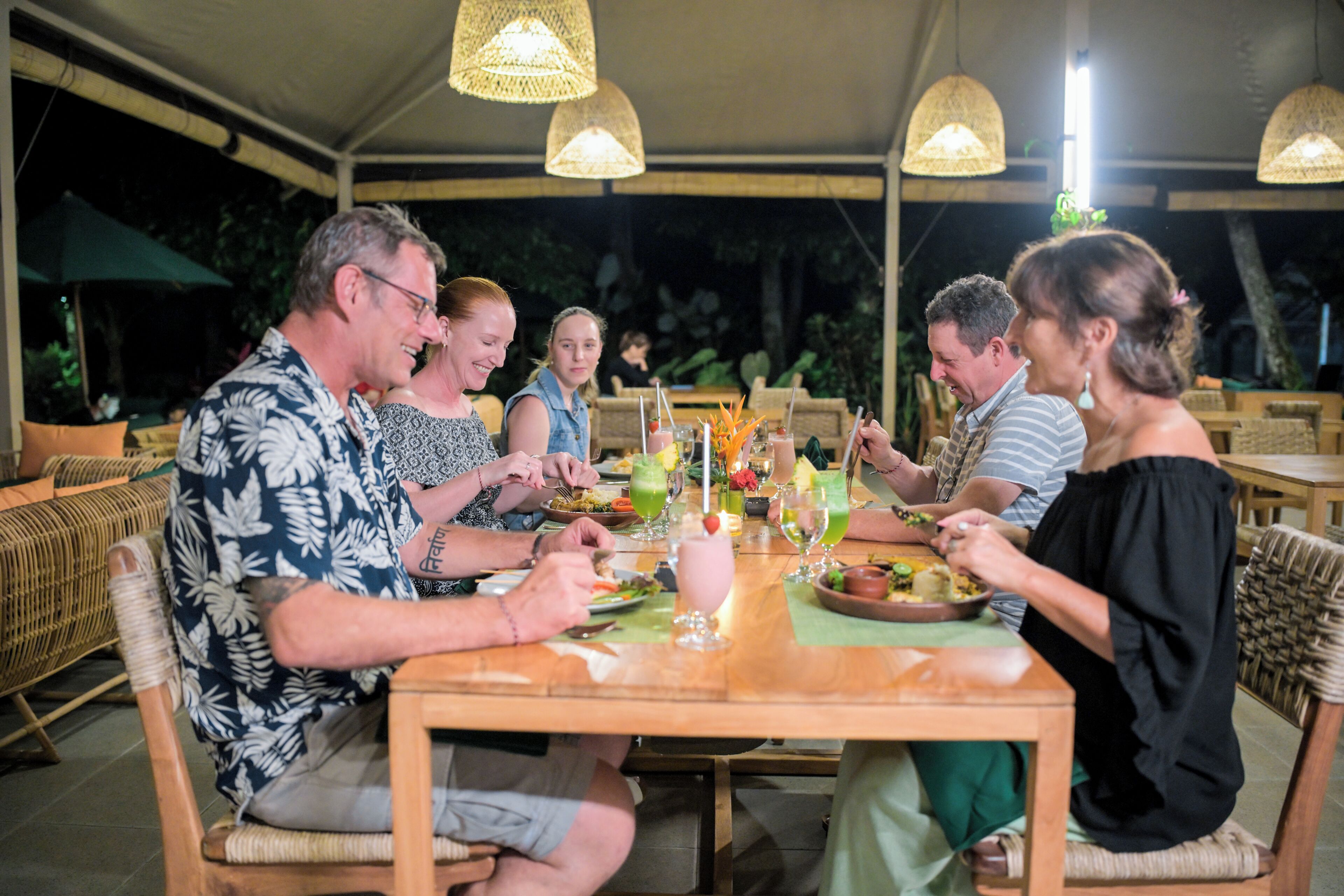 Un groupe de personnes est assis à une table dans un restaurant en plein air, où les meubles sont en osier et des guirlandes lumineuses sont suspendues.