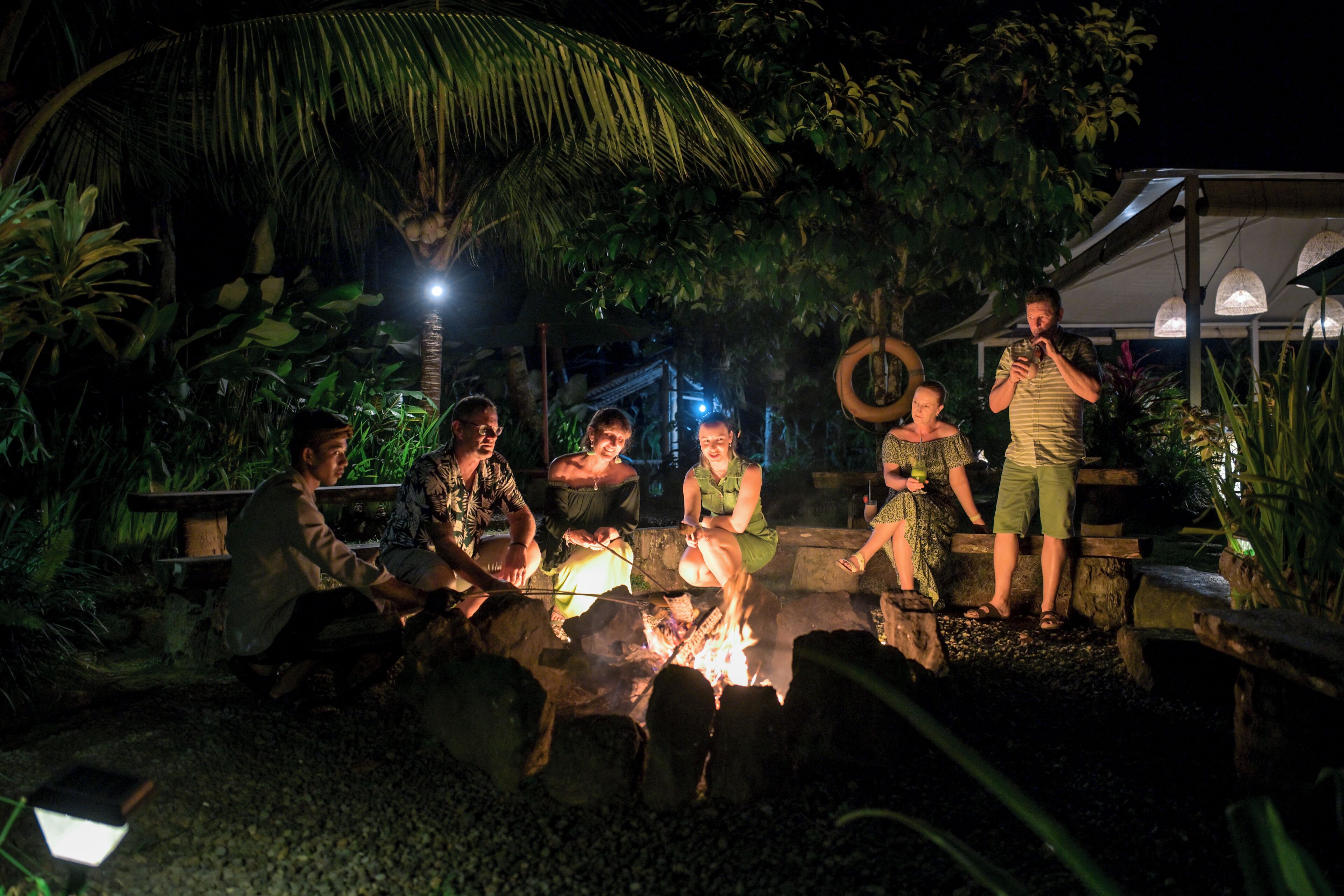 A group of people around a campfire at night, with a thatched roof structure and palm trees in the background.