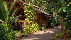A rustic wooden building with a thatched roof, surrounded by lush greenery and a gravel path. - Kaura Bali (Manggis)