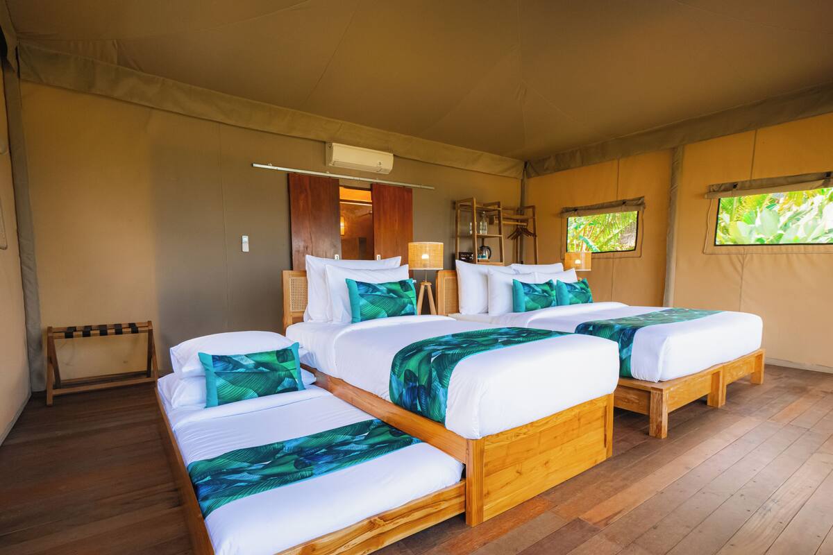 Quarto com duas camas de madeira, um banco de madeira, um ventilador de teto e uma janela com vista para vegetação.