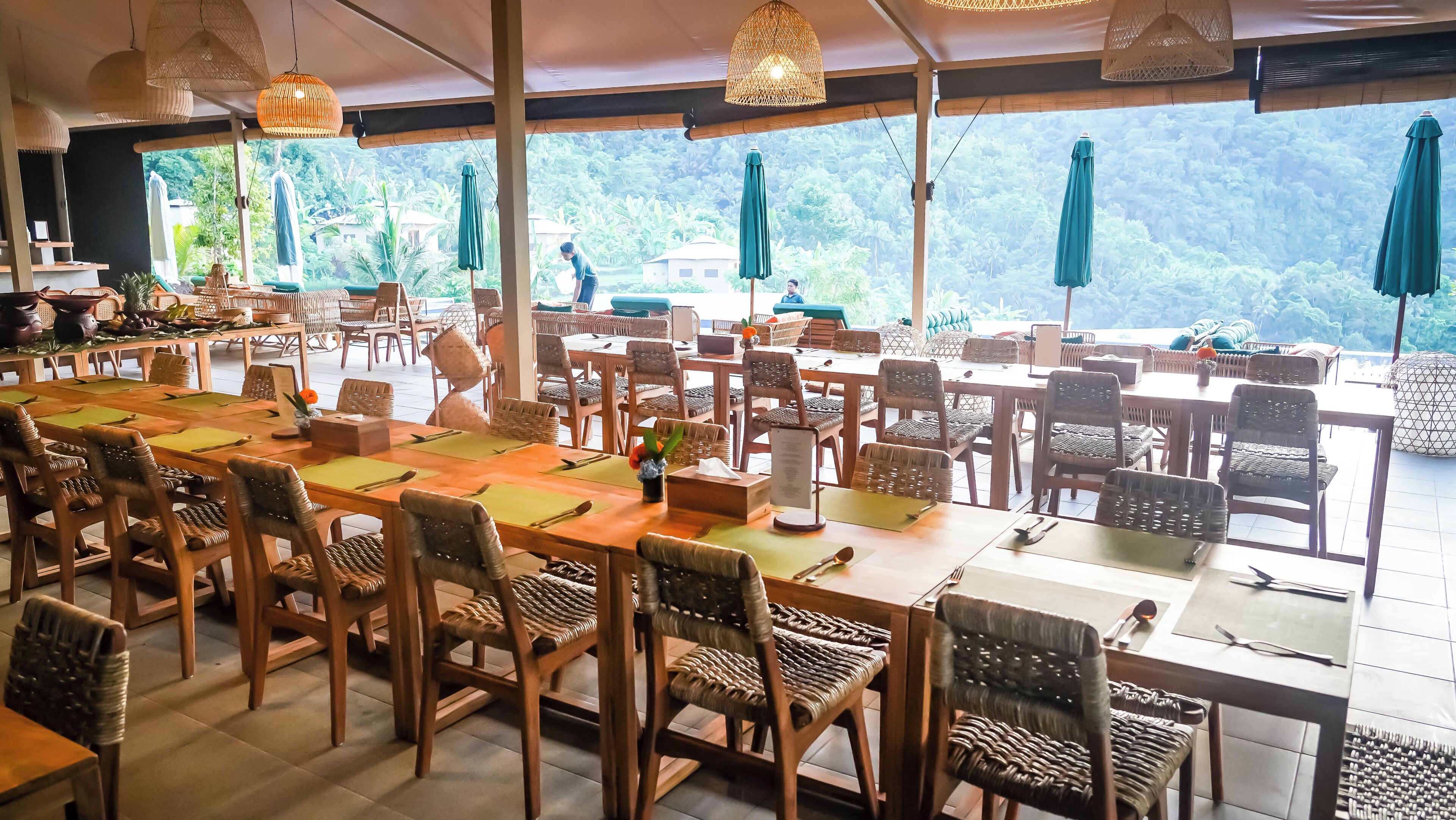 Ein Restaurant mit Holztischen und -stühlen, grünen Sonnenschirmen und Blick auf üppiges Grün.