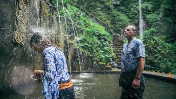A man in a blue patterned shirt and dark pants standing in a water feature with a waterfall, surrounded by greenery.
