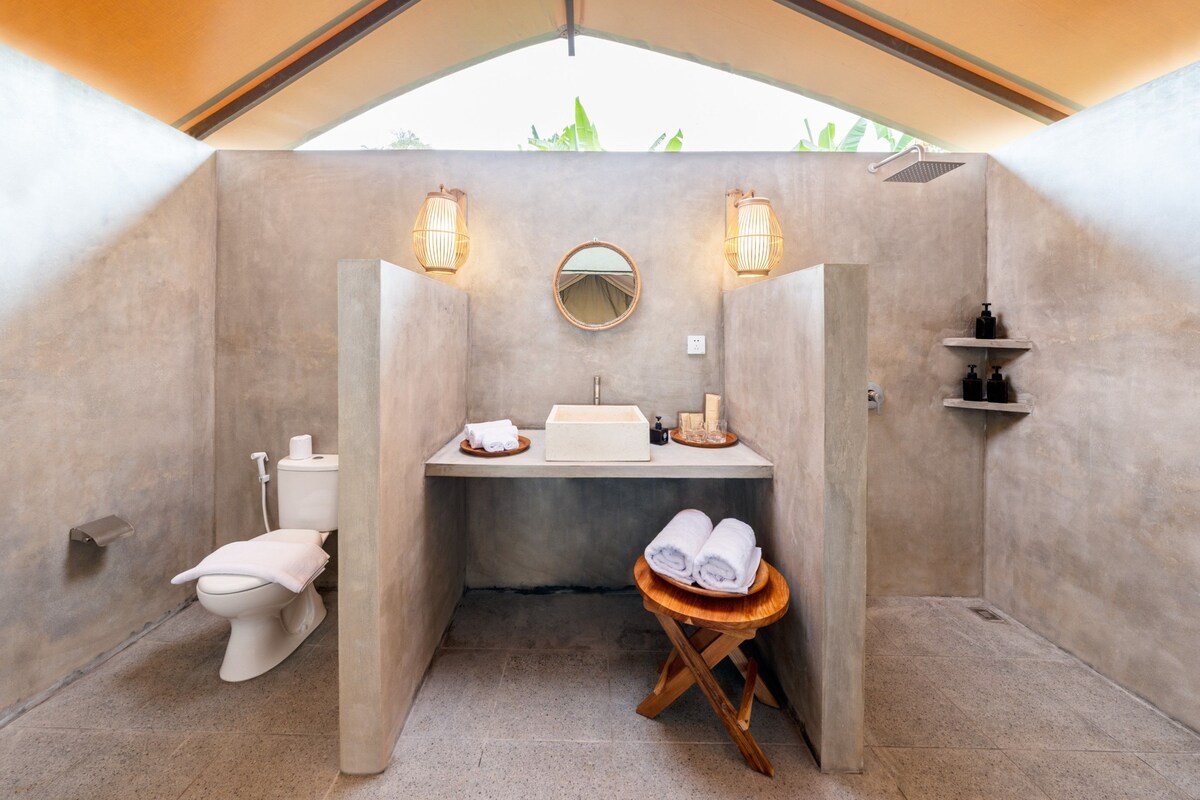 A compact bathroom with a toilet, sink, and shower, featuring a round mirror, two wall-mounted lights, and a small shelf with toiletries.
