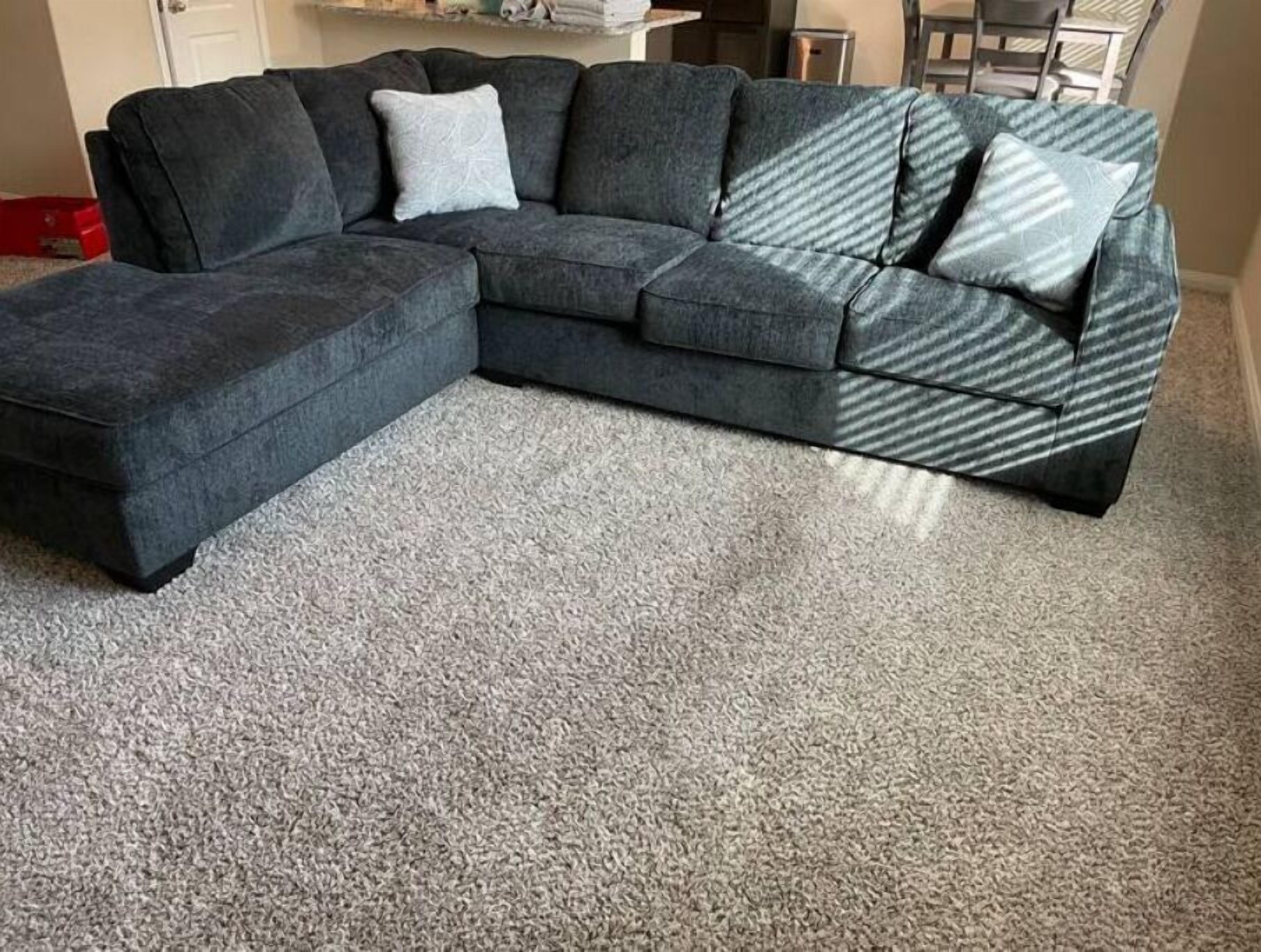 A sectional sofa with a grey and blue patterned accent, a light grey throw pillow, and a grey carpeted floor.