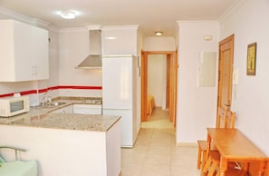 A compact kitchen with white cabinets, a microwave, a stove, and a refrigerator. There is a dining area with a wooden table and chairs. - Apartment Santa Teresa II (Conil de la Frontera)
