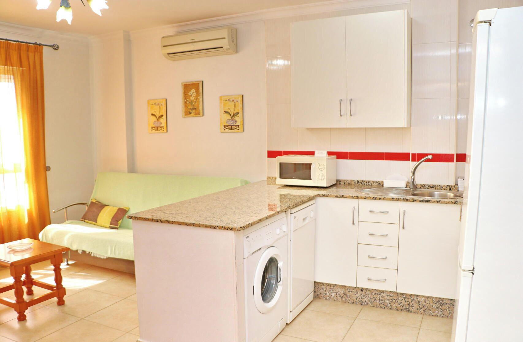 A compact kitchen with a washing machine, a microwave, and a sink. There is a green sofa and a small wooden table in the living area.