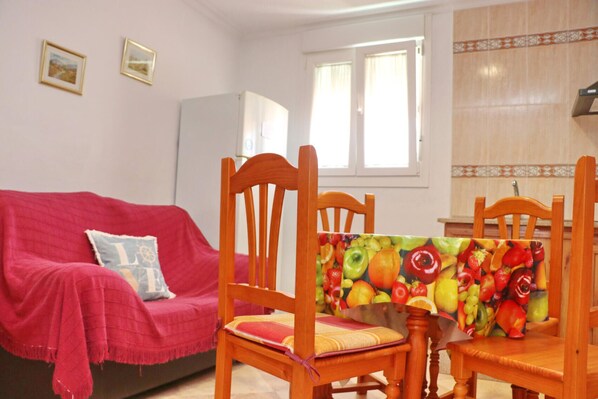 Una sala de estar con un sofá rojo, sillas de madera, una mesa de comedor con mantel estampado de frutas y un refrigerador.