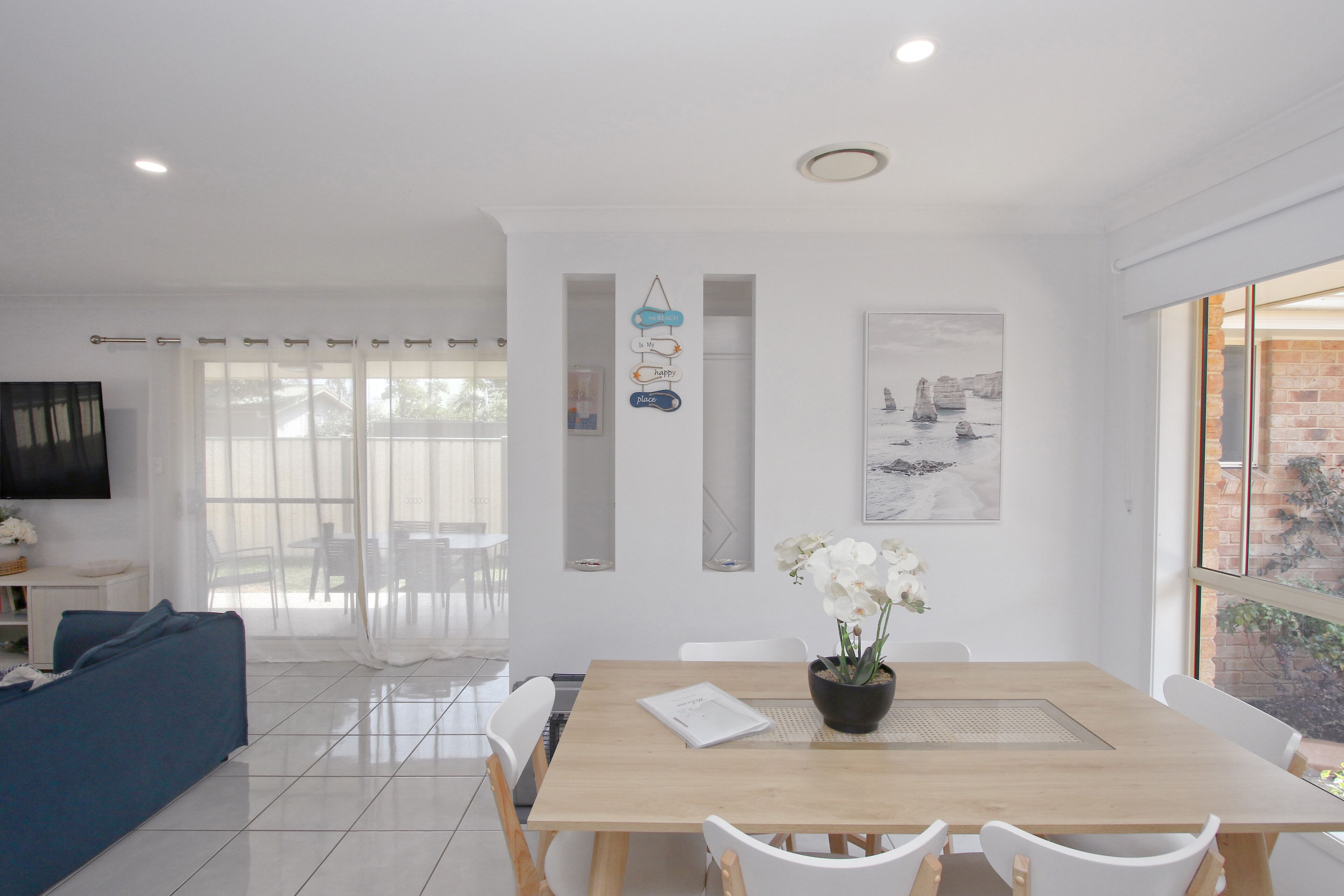 A modern living room with a dining area, white furniture, a flat-screen TV, and a view of the outdoors.