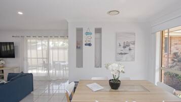 A modern living room with a dining area, white furniture, a flat-screen TV, and a view of the outdoors.