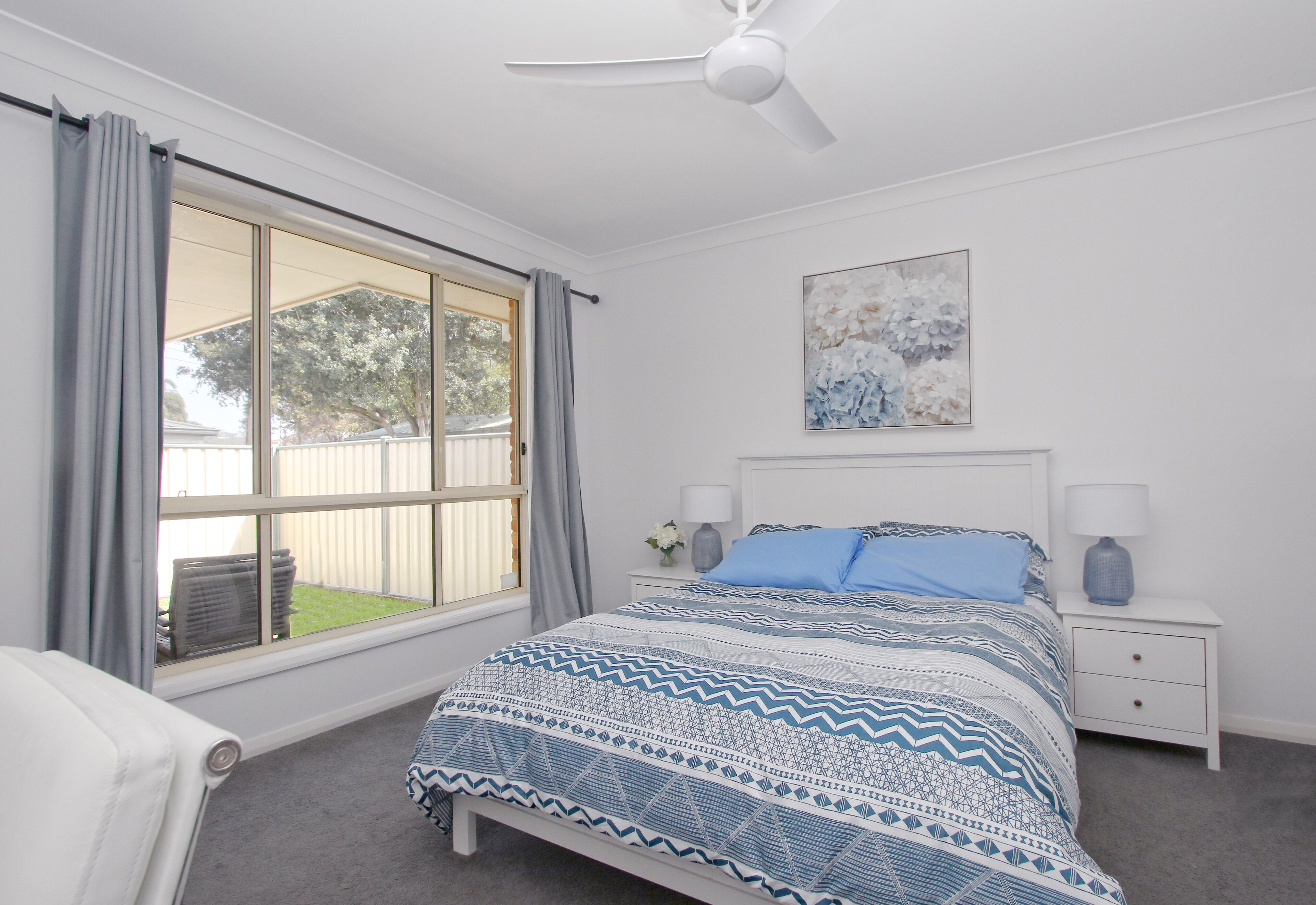A bedroom with a large window, a bed with patterned bedding, a ceiling fan, and a nightstand with a lamp.