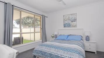 A bedroom with a large window, a bed with patterned bedding, a ceiling fan, and a nightstand with a lamp.