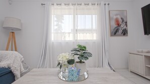 Una habitación con cama, mesita de noche con lámpara, ventana con cortinas, planta en maceta y televisor de pared.