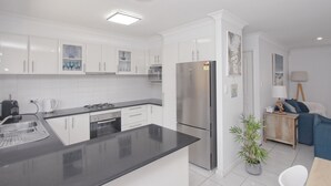 Una cocina moderna con armarios blancos, encimera negra, electrodomésticos de acero inoxidable y un área de comedor con sofá azul y una lámpara.