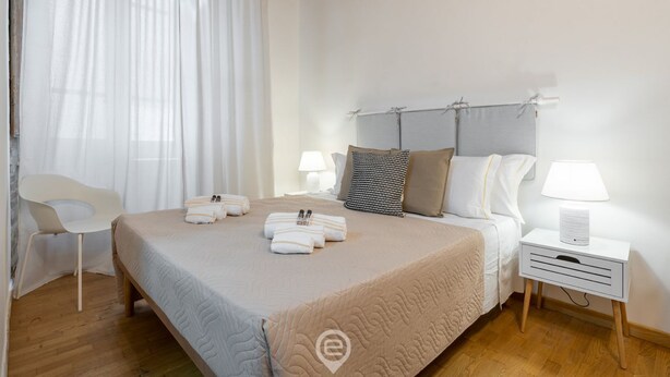 Un dormitorio ordenado con cama, mesitas de noche, una silla y una ventana con cortinas.