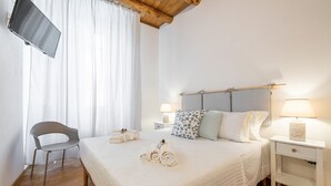 Habitación de hotel con cama, una silla, televisor montado en la pared y ventana con cortinas.