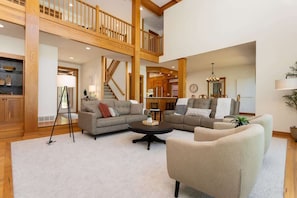A spacious living area with a high ceiling, wooden beams, a staircase, and a view into the kitchen.