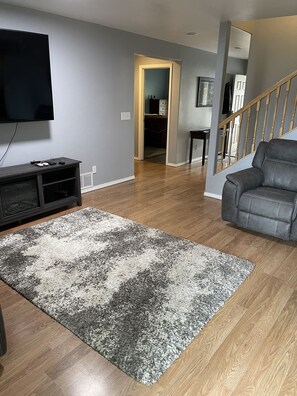 A living room with a flat-screen TV, a dark wooden entertainment unit, a grey and white rug, a grey leather sofa, and a wooden staircase.